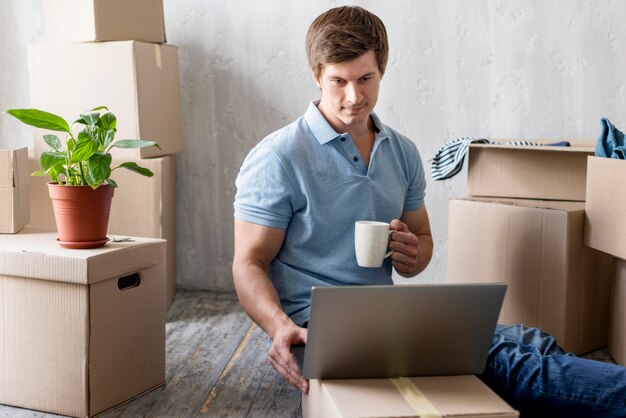 Homem em casa com laptop e caneca organizando caixas para se mudar