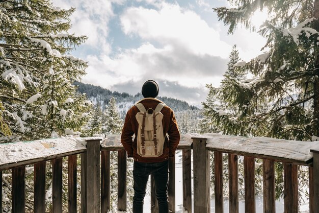 Homem em frente a cercas de madeira, cercado por colinas e florestas cobertas de neve