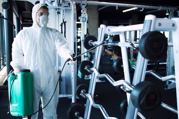 Homem em traje de proteção branco desinfetando e equipamentos de ginástica e pesos para impedir a propagação do vírus corona altamente contagioso