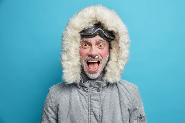 Homem emocional congelado grita alto tem o rosto vermelho coberto de gelo vestido com jaqueta térmica com capuz e óculos de snowboard.