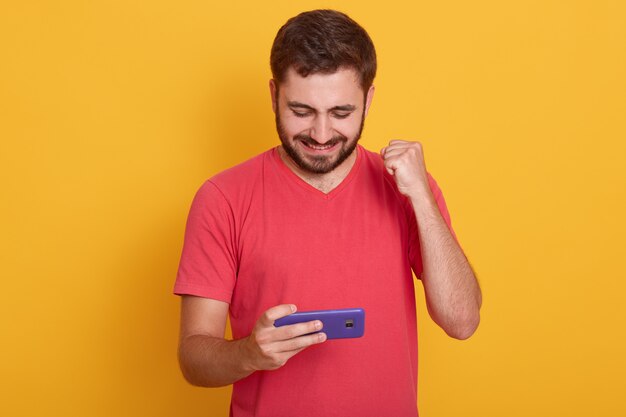 Homem encantado veste camiseta casual vermelha, jogando videogame no celular e punho isolado sobre o estúdio amarelo