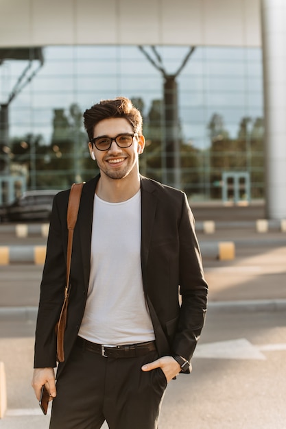 Foto grátis homem encantador de terno preto, camiseta branca e óculos, sorri amplamente e olha para a câmera