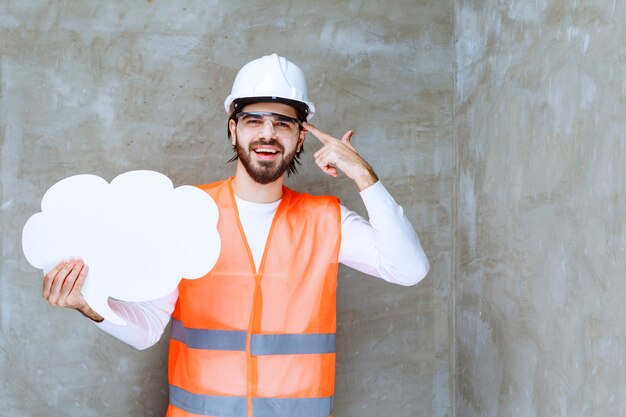 Homem engenheiro com capacete branco e óculos de proteção segurando um quadro de informações e parece confuso e apavorado.