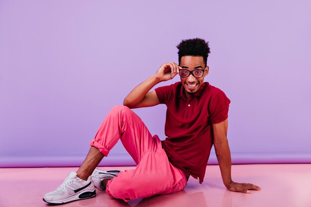 Homem engraçado africano com cabelo curto, sentado no chão. Retrato de um cara inspirado de óculos posando de brincadeira.