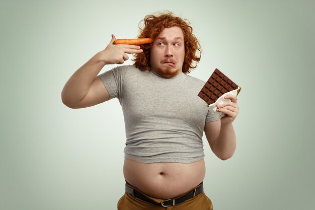 Homem engraçado com excesso de peso segurando uma barra de chocolate em uma das mãos e uma cenoura na têmpora como uma arma