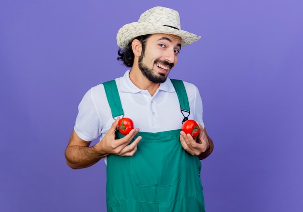 Homem engraçado jovem barbudo jardineiro vestindo macacão e chapéu segurando tomates frescos olhando para frente sorrindo alegremente em pé sobre a parede azul