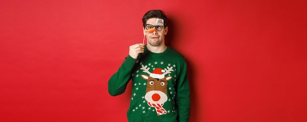 Homem engraçado no suéter de Natal e máscara de festa, comemorando as férias de inverno, mostrando caretas ...