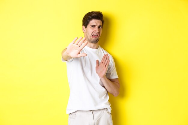 Homem enojado se recusando, fazendo uma careta de antipatia e aversão, implorando para parar, em pé com uma camiseta branca contra um fundo amarelo.