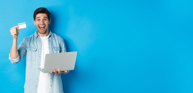 Foto grátis homem entusiasmado faz compras on-line mostrando cartão de crédito e segurando laptop comprando na internet em pé sobre azul