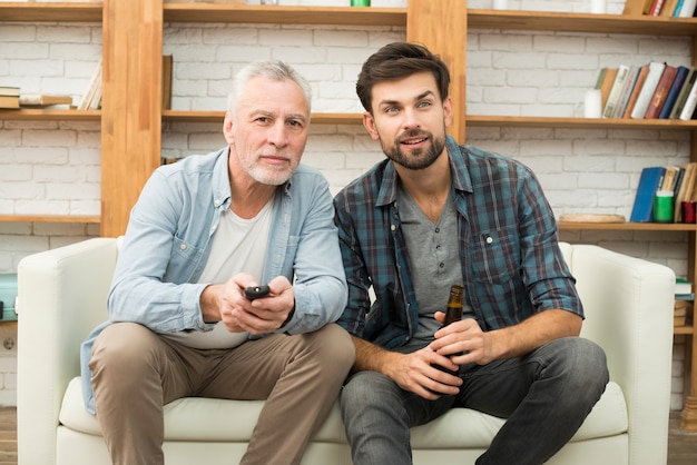 Homem envelhecido com controle remoto e cara jovem com garrafa assistindo tv no sofá