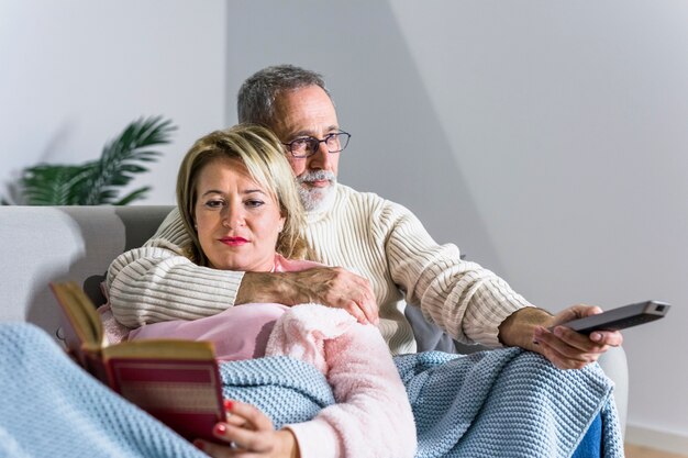 Homem envelhecido, com, controle remoto tv, observar, tv, e, leitura mulher, livro, ligado, sofá