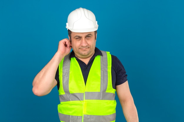 Homem envelhecido médio vestindo colete amarelo de construção e capacete de segurança tocando sua expressão suspeita de orelha sobre parede azul isolada