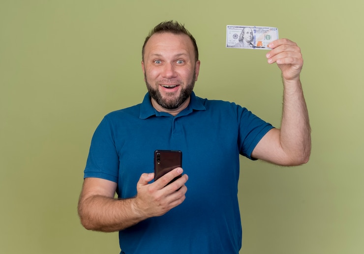 Foto grátis homem eslavo adulto alegre levantando dinheiro e segurando um telefone celular