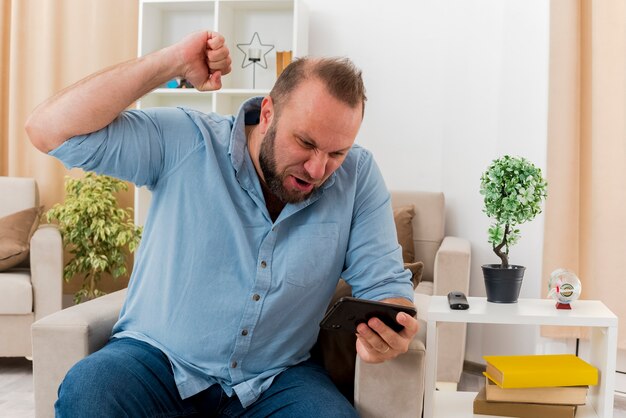 Homem eslavo adulto alegre sentado na poltrona, levantando o punho e olhando para o telefone dentro da sala de estar