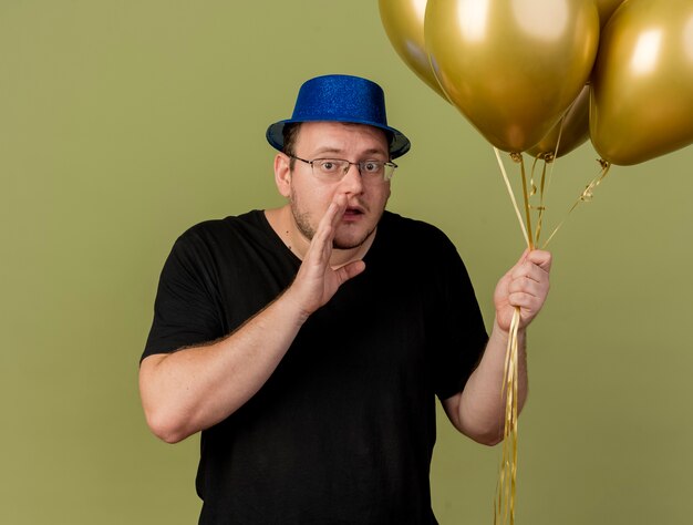 Homem eslavo adulto ansioso com óculos óticos e chapéu de festa azul mantém a mão perto da boca e segura balões de hélio
