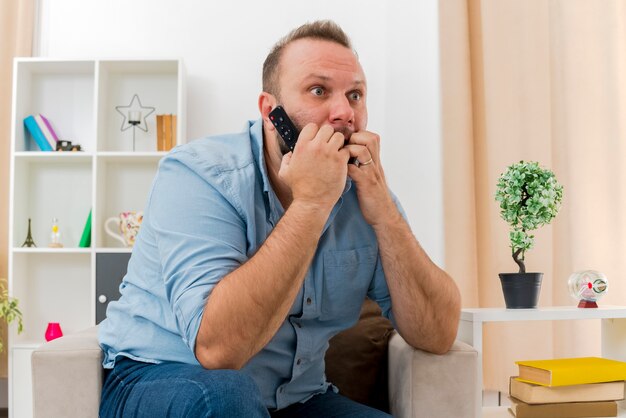 Homem eslavo adulto ansioso sentado na poltrona segurando o controle remoto da TV, mordendo os dedos olhando para o lado dentro da sala de estar