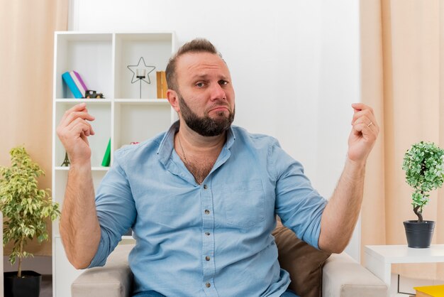 Homem eslavo adulto chateado sentado na poltrona gesticulando sinal de dinheiro com as duas mãos olhando para a câmera dentro da sala de estar