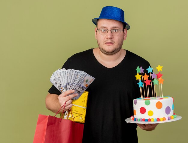 Homem eslavo adulto chocado com óculos ópticos e chapéu de festa azul segurando uma caixa de dinheiro, sacola de papel e bolo de aniversário