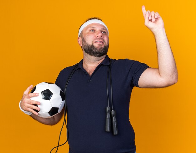 Homem eslavo adulto e esportivo satisfeito com pular corda em volta do pescoço, usando bandana e pulseiras segurando uma bola e apontando para cima, isolado na parede laranja com espaço de cópia