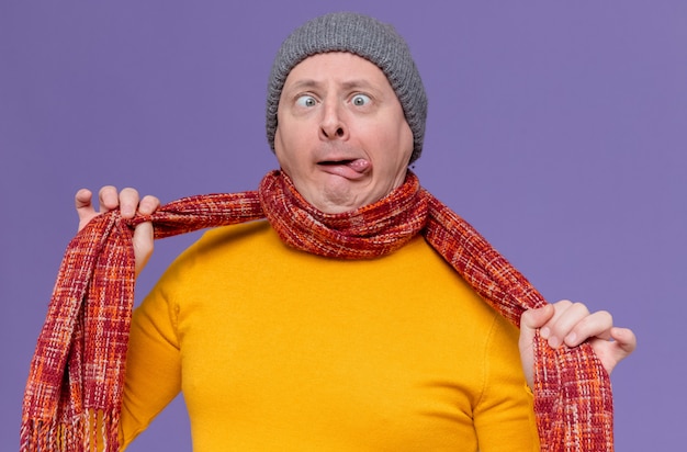 Foto grátis homem eslavo adulto engraçado com chapéu de inverno e cachecol no pescoço, metendo a língua para fora e se sufocando com o cachecol