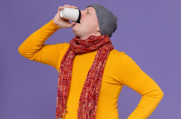 Homem eslavo adulto insatisfeito com chapéu de inverno e lenço no pescoço, bebendo em copo de papel
