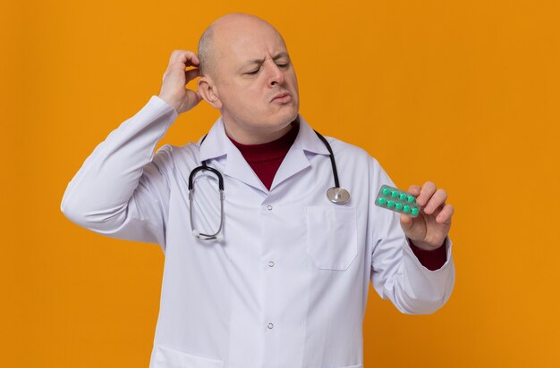 Homem eslavo adulto pensativo em uniforme de médico com estetoscópio segurando e olhando para a embalagem de remédio