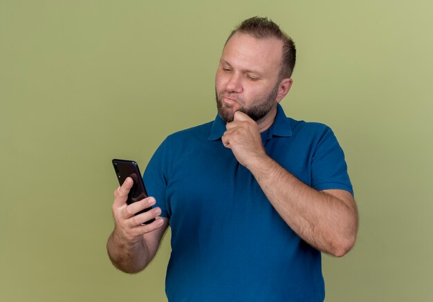 Homem eslavo adulto pensativo segurando e olhando para o celular e tocando o queixo