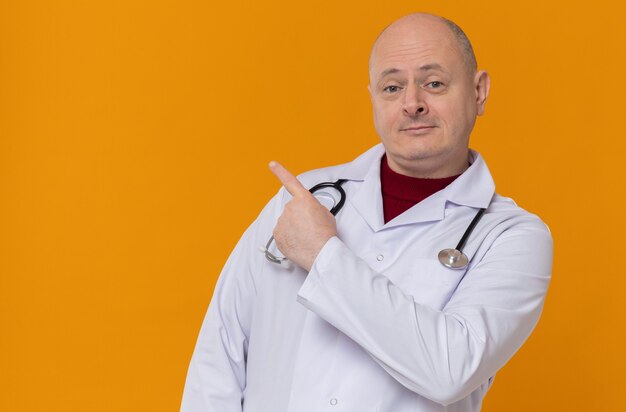 Homem eslavo adulto satisfeito com uniforme de médico e estetoscópio apontando para o lado