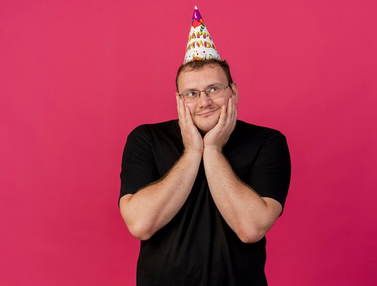 Foto grátis homem eslavo adulto satisfeito usando óculos óticos e boné de aniversário colocando as mãos no rosto