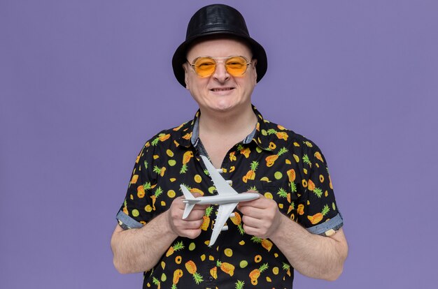 Homem eslavo adulto sorridente com cartola preta e óculos escuros segurando o modelo do avião