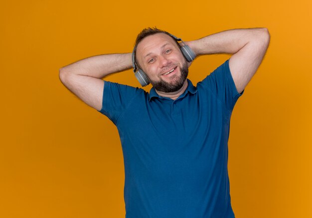 Homem eslavo adulto sorridente usando fones de ouvido, olhando para ouvir música e mantendo as mãos atrás da cabeça