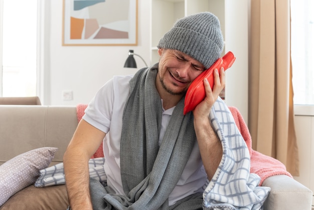 Foto grátis homem eslavo doente a chorar com um lenço no pescoço e chapéu de inverno segurando uma garrafa de água quente sentado no sofá da sala