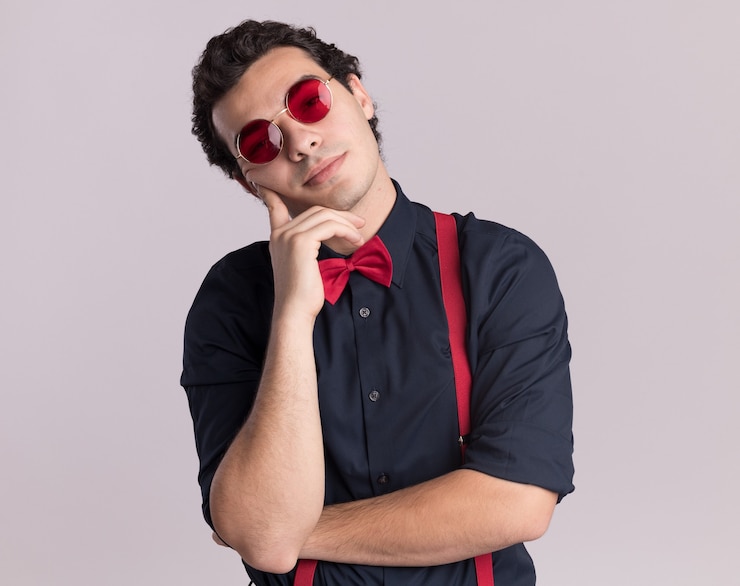 Foto grátis homem estiloso com gravata borboleta usando óculos e suspensórios, olhando para a frente com expressão pensativa e pensando em pé sobre uma parede branca
