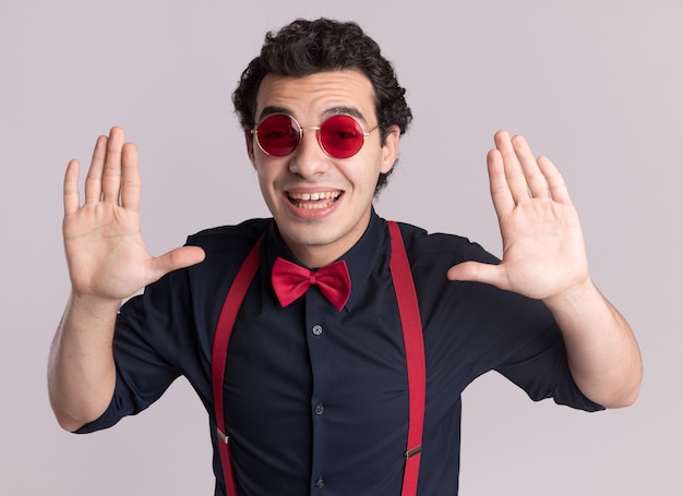 Homem estiloso com gravata borboleta usando óculos e suspensórios, olhando para a frente, sorrindo amplamente com os braços erguidos em pé sobre uma parede branca