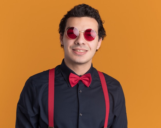 Foto grátis homem estiloso com gravata borboleta usando óculos e suspensórios, olhando para a frente, sorrindo feliz e positivo em pé sobre a parede laranja