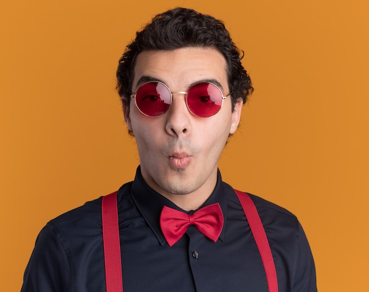 Foto grátis homem estiloso com gravata borboleta usando óculos e suspensórios olhando para a frente surpreso em pé sobre a parede laranja