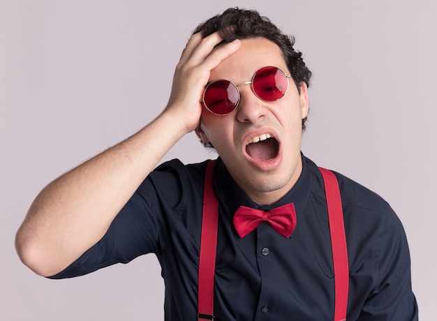 Homem estiloso com gravata borboleta usando óculos e suspensórios olhando para frente confuso com a mão na cabeça gritando com uma expressão irritada em pé sobre uma parede branca