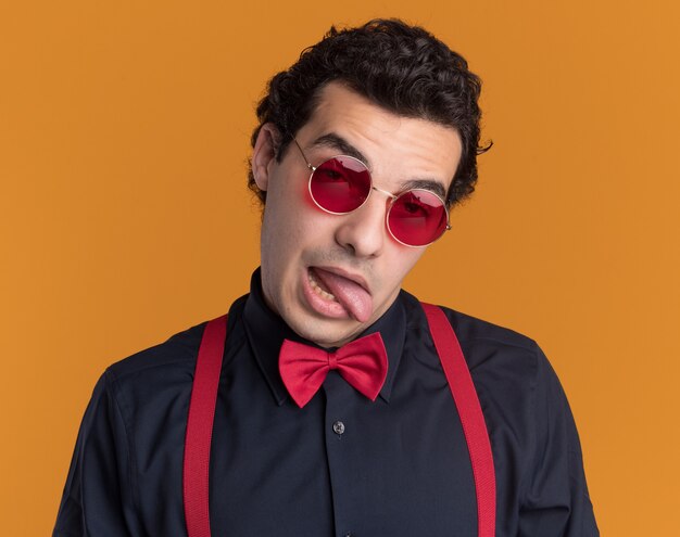 Homem estiloso com gravata borboleta usando óculos e suspensórios olhando para frente fazendo careta e mostrando a língua em pé sobre a parede laranja