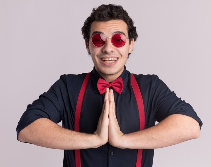 Foto grátis homem estiloso com gravata borboleta usando óculos e suspensórios segurando as palmas das mãos juntas como um gesto namastê feliz e alegre sorrindo em pé sobre uma parede branca