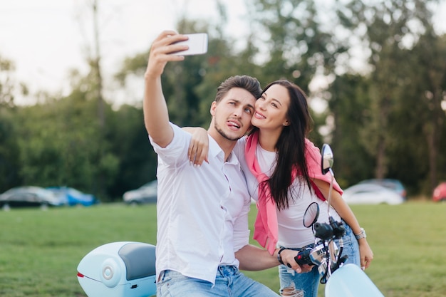 Homem estiloso na scooter fazendo caretas engraçadas enquanto tira fotos com uma mulher elegante de cabelos escuros