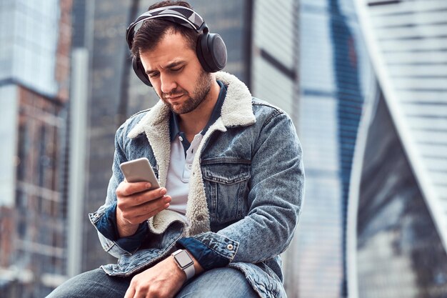 Homem estiloso vestindo jaqueta jeans ouvindo música e usando smartphone sentado na frente de arranha-céus na cidade de Moscou na manhã nublada.