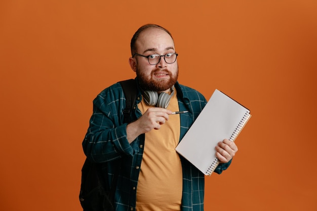 Homem estudante em roupas casuais usando óculos com fones de ouvido e mochila segurando caderno e caneta olhando para a câmera confuso em pé sobre fundo laranja