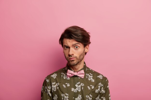 Homem europeu confuso com penteado moderno olha diretamente para a câmera, usa camisa verde com gravata borboleta, isolado sobre uma parede rosada