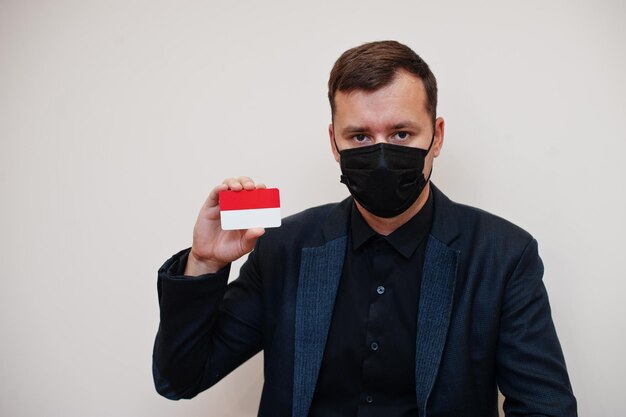 Homem europeu usa formal preto e protege máscara facial segura cartão de bandeira de Mônaco isolado em fundo branco Europa coronavírus Covid conceito de país