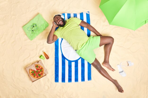 homem exclama em voz alta gosta de ouvir música com fones de ouvido usa camiseta verde e bermuda faz um lanche saboroso deitado em toalhas listradas na praia.