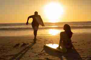 Foto grátis homem, executando, com, surfboard, enquanto, mulher relaxando, praia, durante, pôr do sol