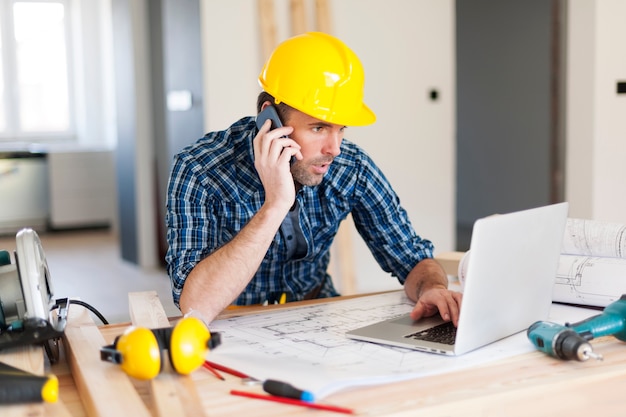 Homem falando no celular e usando laptop na construção