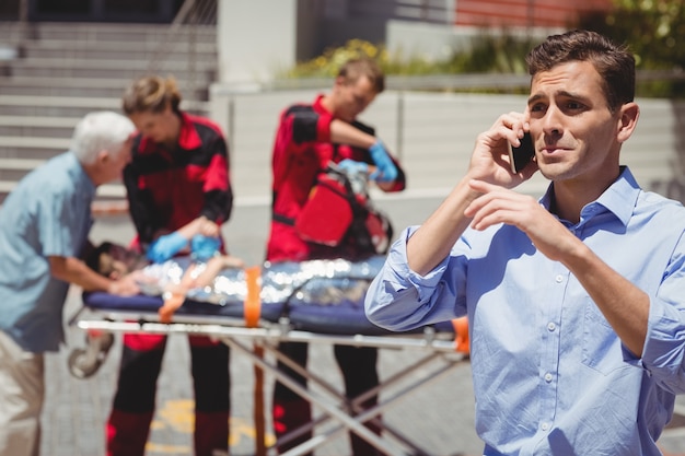 Homem falando no telefone celular e paramédicos examinando menino ferido no fundo