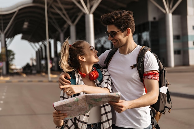 Homem feliz abraça sua namorada loira atraente viajantes com mochilas seguram mapa e falam mulher bonita em sorrisos de camisa xadrez e óculos de sol