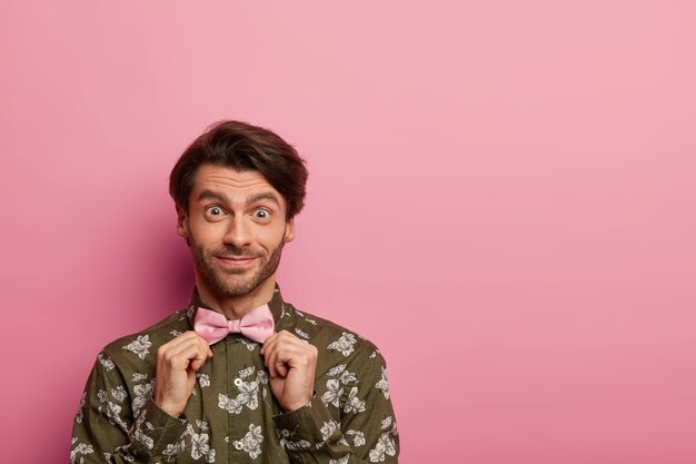 Homem feliz com a barba por fazer ajusta a gravata borboleta, usa uma camisa da moda com estampa de flores e se prepara para o primeiro encontro